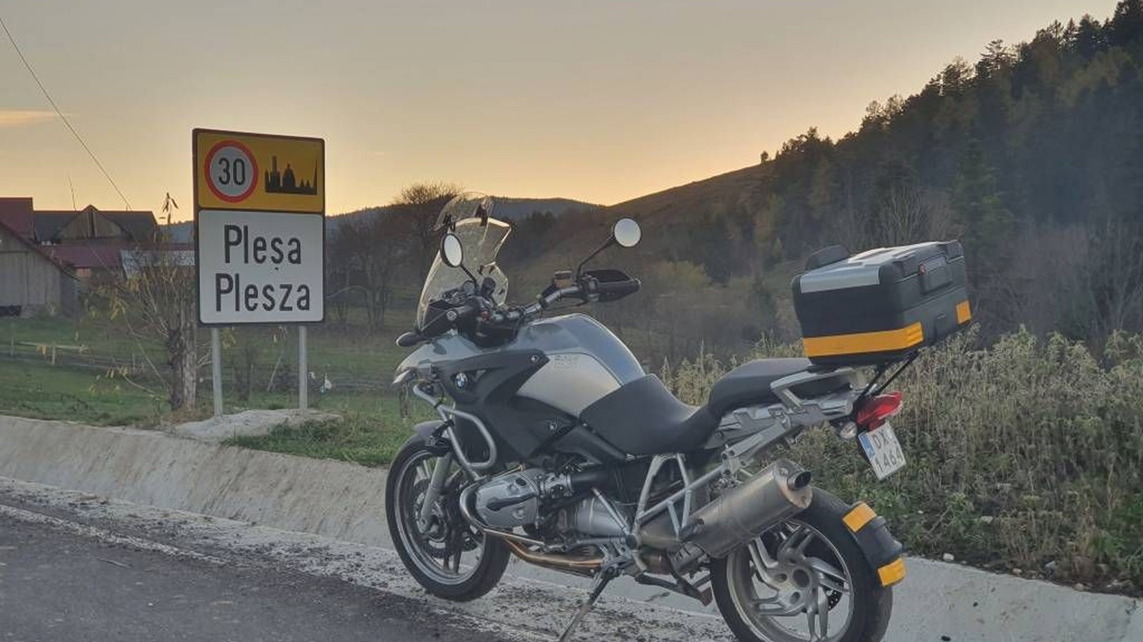 Motocicletă lângă tăblița cu numele satului în două limbi, Pleșa și Plesza, la apus