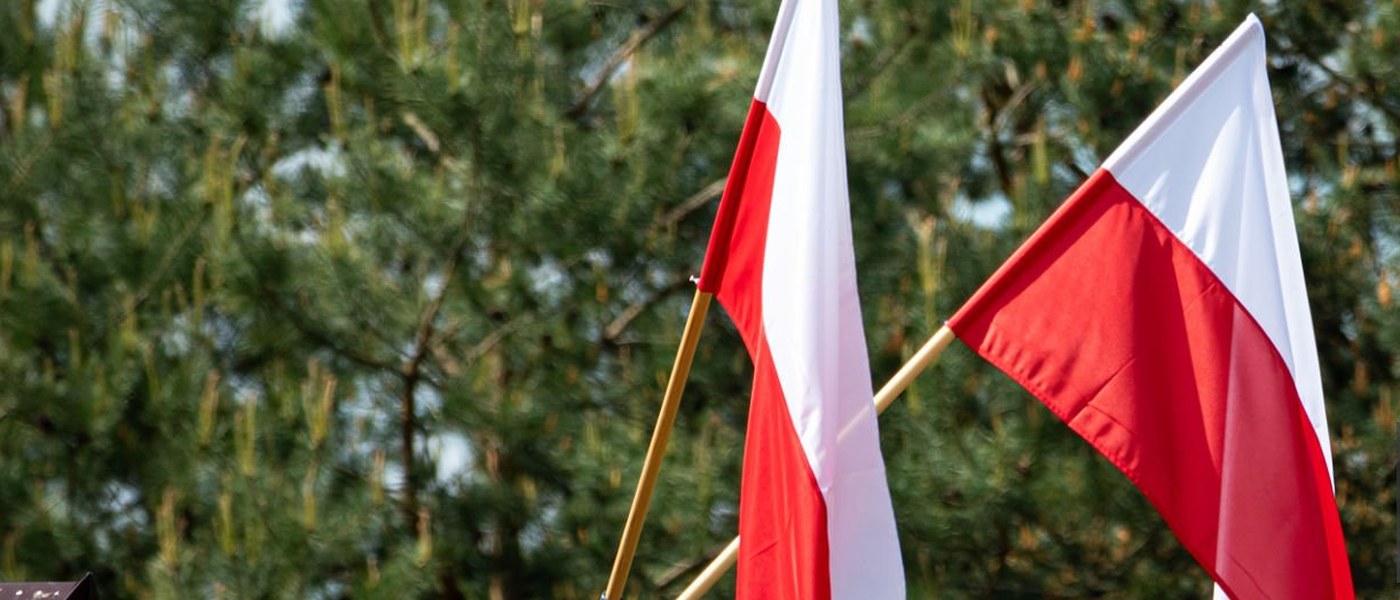 Dwie polskie flagi na drewnianym słupie, na tle sosnowego lasu